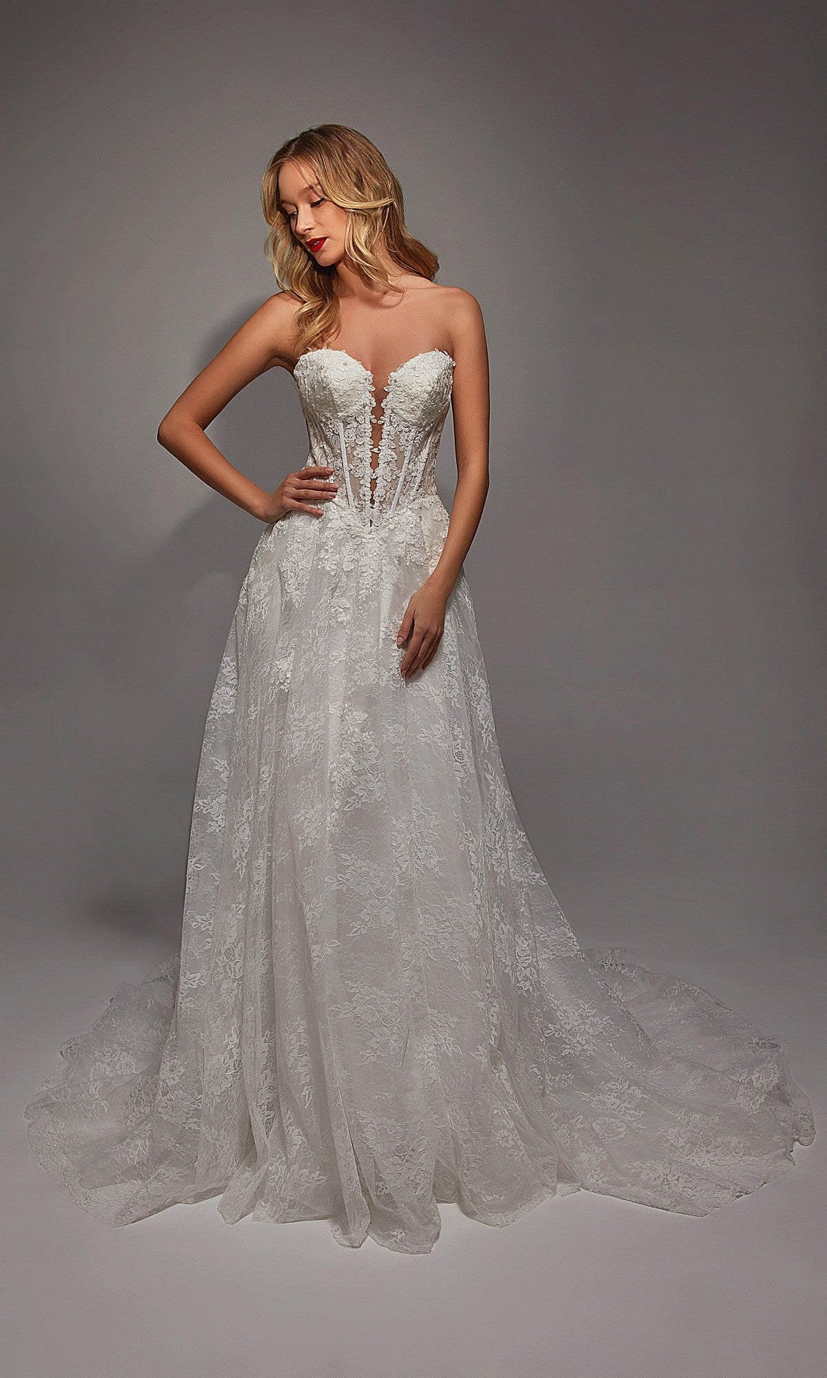 A woman poses against a plain gray background in the Alyce 7154 long white dress, featuring a strapless sheer corset bodice, lace detailing, and a flowing train.